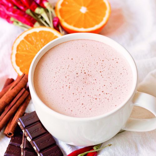 a birds eye view of a froth mug of hot chcoclate ona light surface surrounded by orange slices, cinnamon sticks, chocolate bars, and red chiles