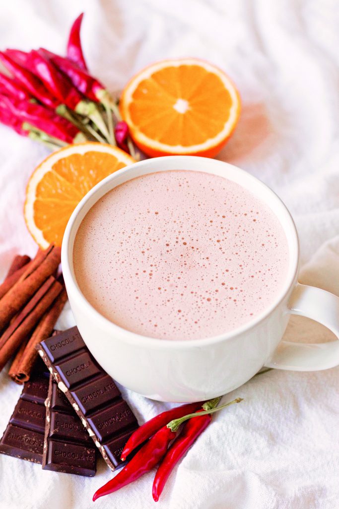 a birds eye view of a froth mug of hot chcoclate ona light surface surrounded by orange slices, cinnamon sticks, chocolate bars, and red chiles