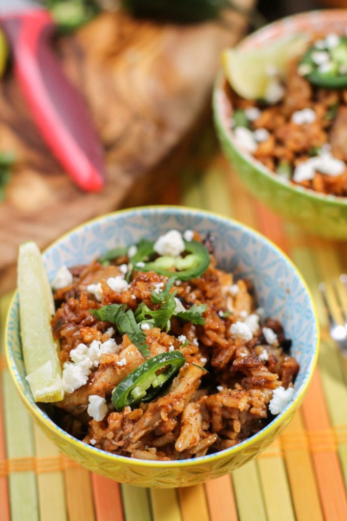 festive colorful bowl filled with a tomato rice and chicken dish topped with fresh lime wedge, chopped cilantro, and cotija cheese