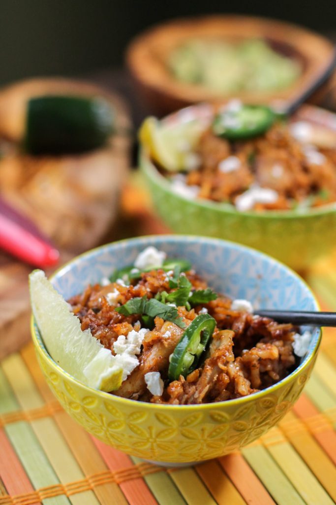 two colorful bowl filled with tomaot rice and tender chicken chunks topped with cilantro, lime, and feta atop a lime, tangerine and yellow colored bamboo mat