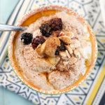 an overhead shot of a orange patterned bowl filled with a creamy porridge topped with blackberries, browned butter, cinnamon, and sliced almonds atop a blue washed wooden surface