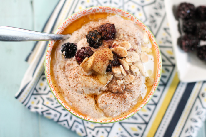 an overhead shot of a orange patterned bowl filled with a creamy porridge topped with blackberries, browned butter, cinnamon, and sliced almonds atop a blue washed wooden surface