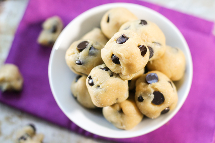  a bowl of raw chocolate chip studded cookie dough balls stacked in a white bowl atop a purple cloth from overhead.