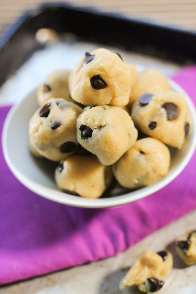 o bake cookie dough balls studded with chocolate chips from above on top of a bright purple cloth and weathered cookie sheet