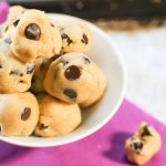 overhead shot of raw chocolate chip cookie dough balls in a white bowl atop a purple fabric and stainless steel cookie sheet