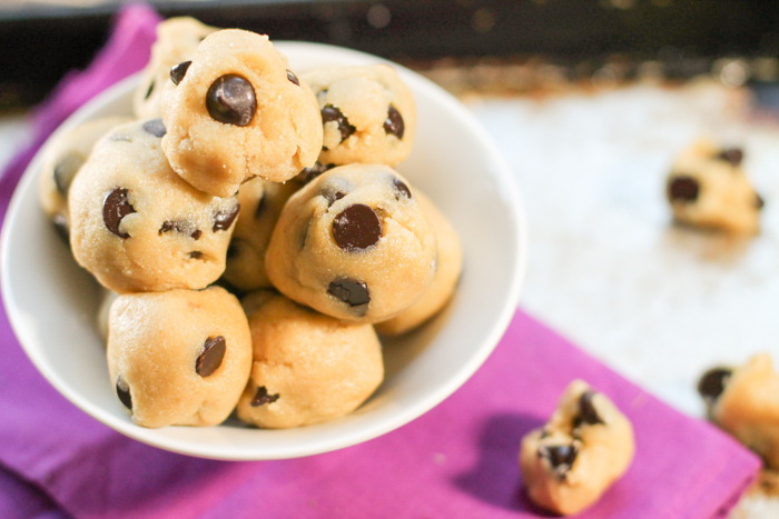 overhead shot of raw chocolate chip cookie dough balls in a white bowl atop a purple fabric and stainless steel cookie sheet