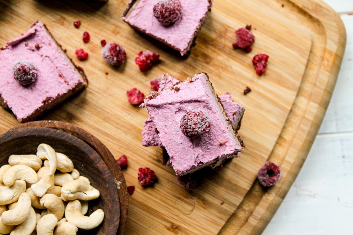 overhead looking down on three raspberry cheescake squares garnished wth fresh raspberries and a bowl of raw cashews