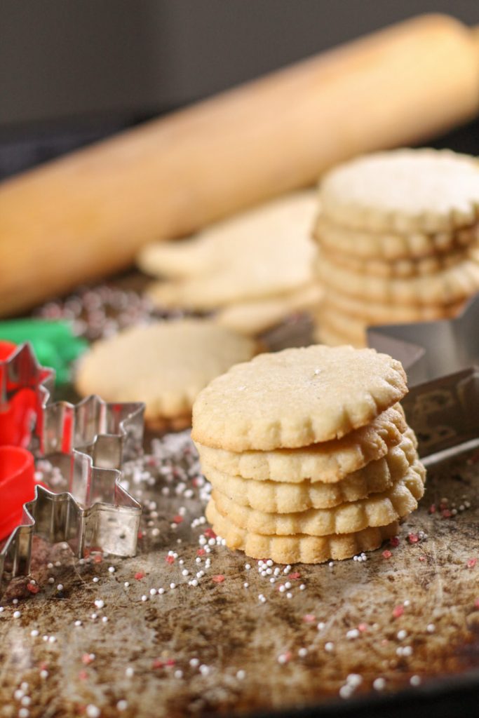 stacked cut out sugar cookies with a rolling pin and festive cookie cutters atop a aged cookie sheet