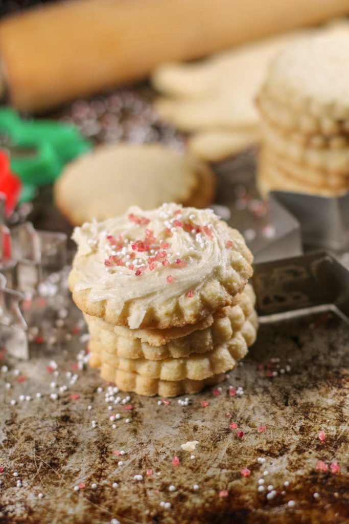 stacked gluten free suagr cookies topped with a creamy frosting and natural red sprinkles atop of antiqued cookie sheet with christmas cookie cutters in background