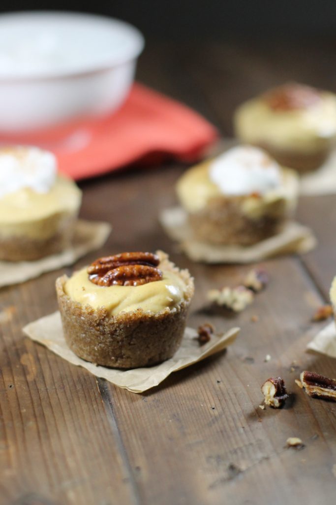 mini pumpkin cheesecakes with a raw nut crust topped with a candied pecan and arranged on a dark wooden table