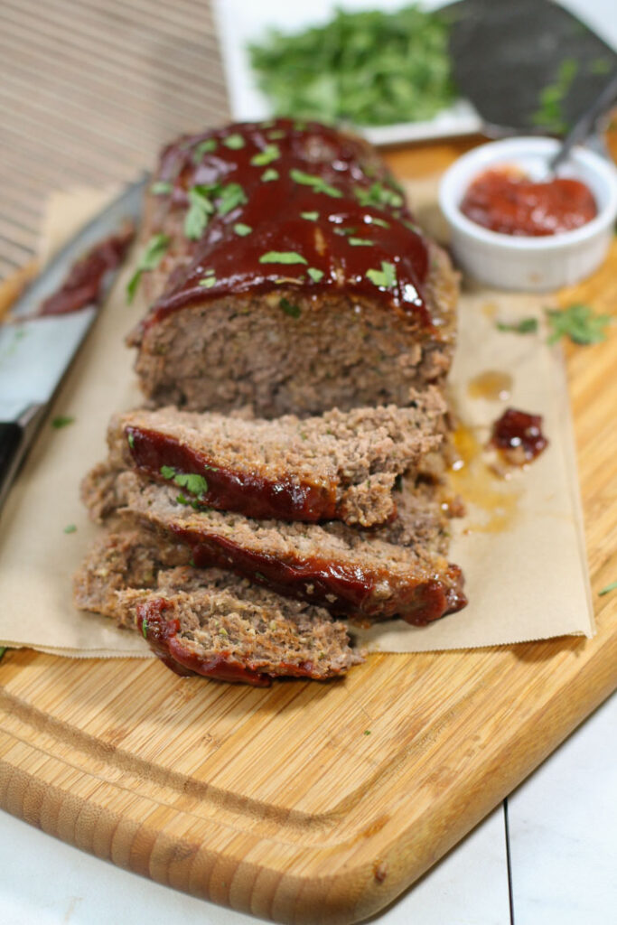 sliced meatloaf topped with a shiny red sauce atop a wooden cutting board and a garnish of chopped parsley.
