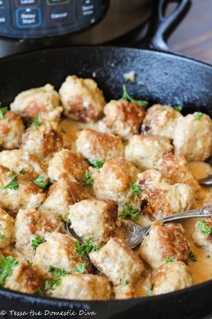 a cast iron skillet filled with browned turkey meatballs slathered in a cream sauce with a garnish of fresh parsley and an instant pot in the back ground