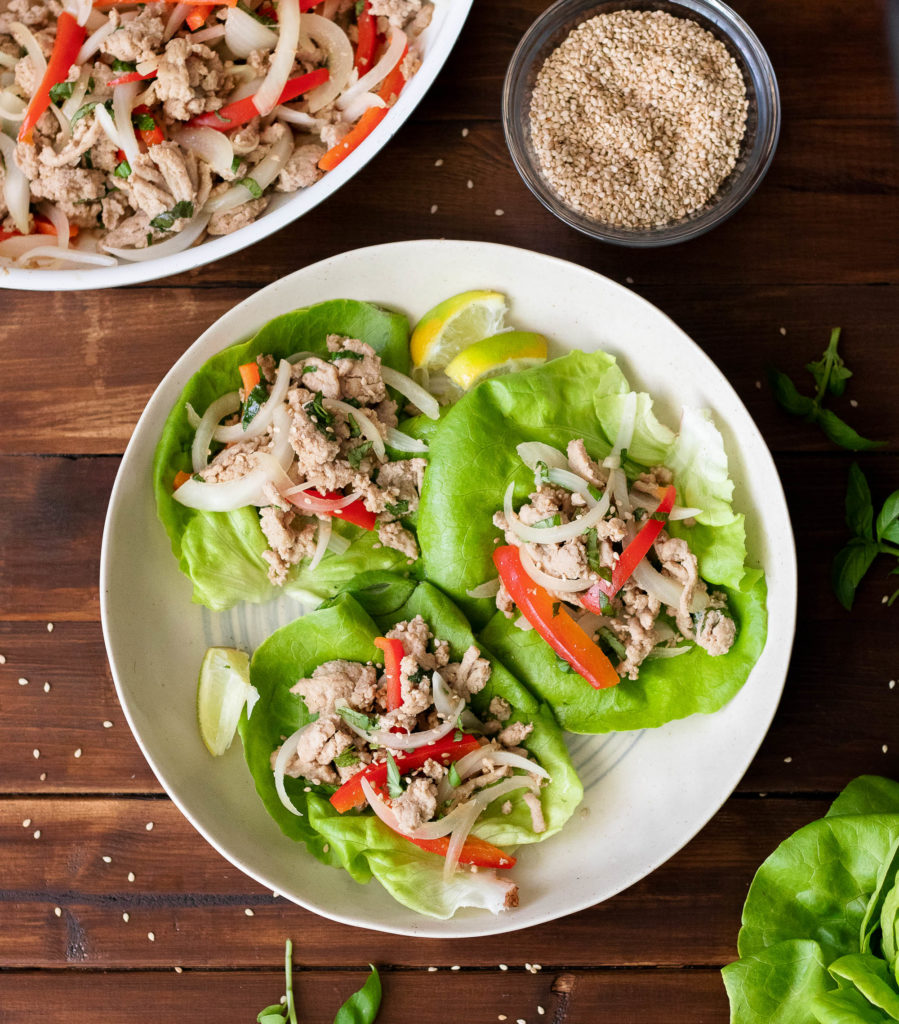 birsdeye view of a two white bowls on a dark wood surface filled with single butter lettuce leaves toped with ground turkey, cooked onion, cooked red peppers, and a garnish of fresh basil and sesame seeds