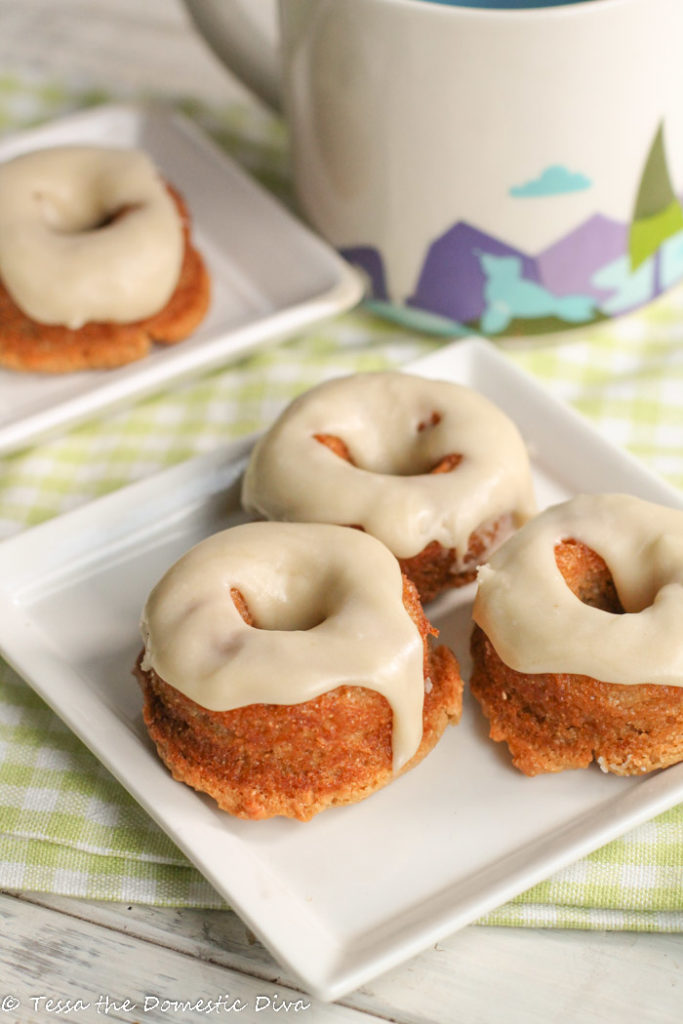 three vanilla cake donuts drizzled with vanilla glaze atop a square white plate with coffee mug in background