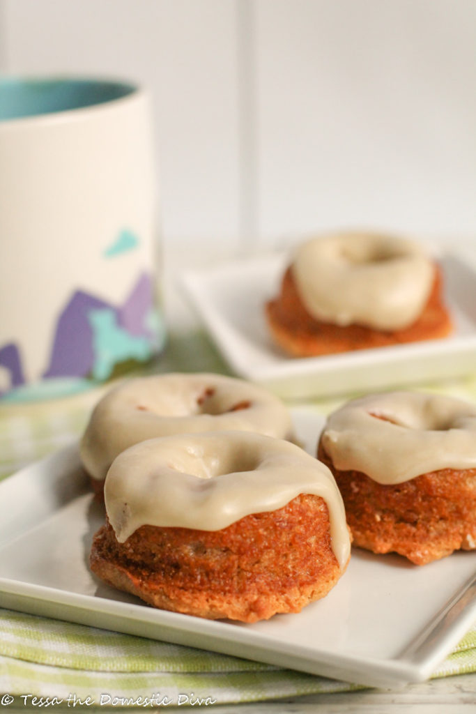 baked vanilla donuts with a smooth vanilla glaze arranged on a sqaure white plate with a colorful coffee mug in the background