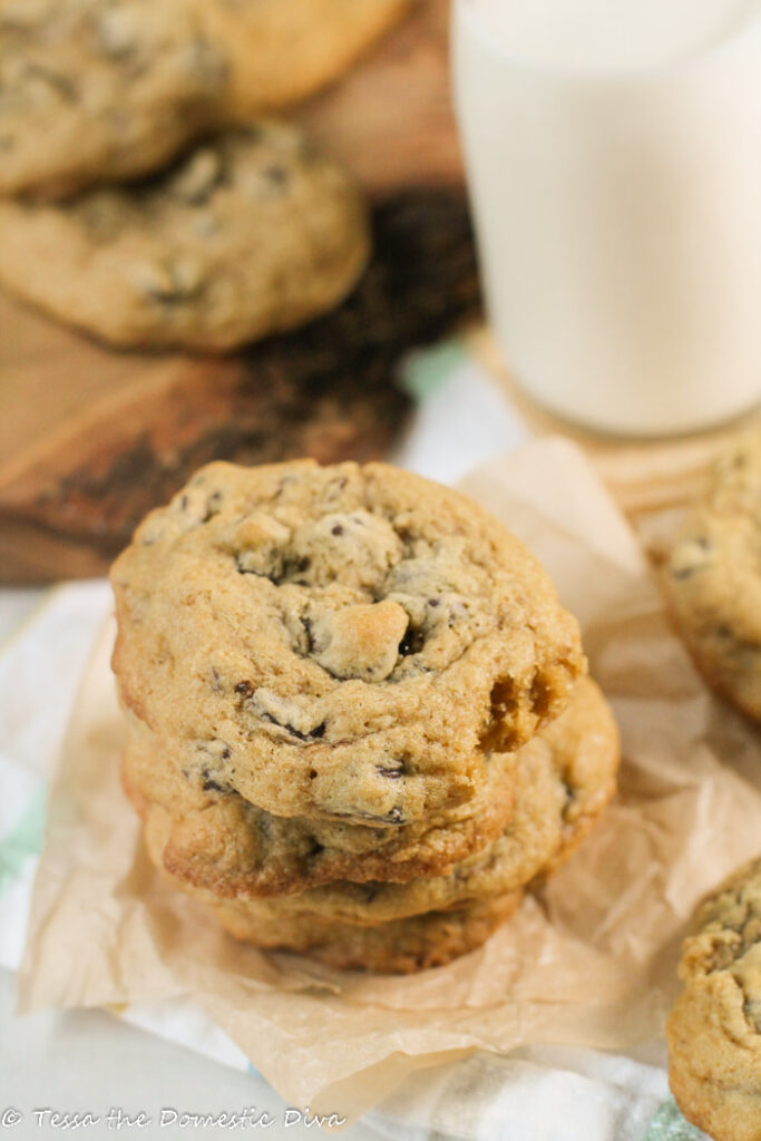 stacked chococlate chip cookies on a square of unbleached parchement paper with addtional arranged in back ground served with a glass of milk