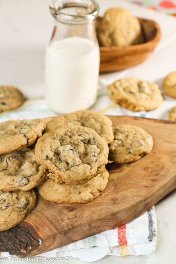 chewy chocolate chip cookies arranged on an olive wood board with a vintage milk glass on a white surface