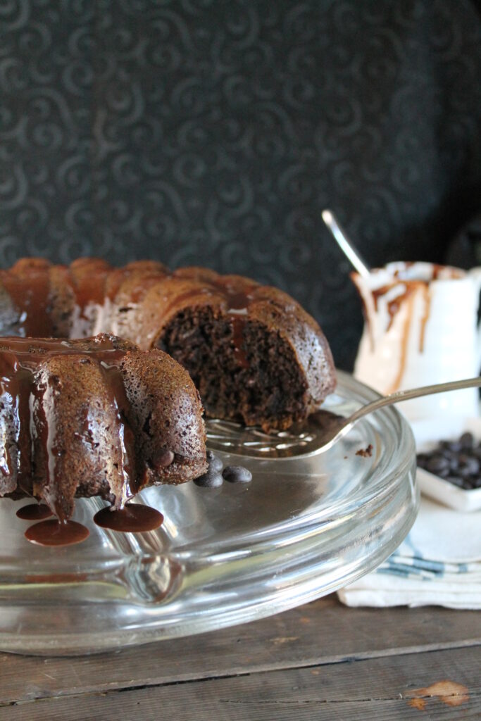 a clear glass cake plate topped with a dark chocolate bundt cake with a a few slices removed and a melted chocolate drizzle