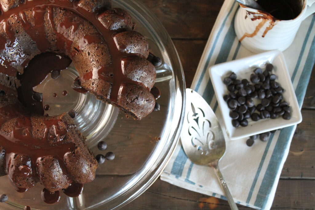 overhead view of a partially sliced chocolate bundt cake on a clear glass cake plate with chocolate chips and melted chocolate drizzle