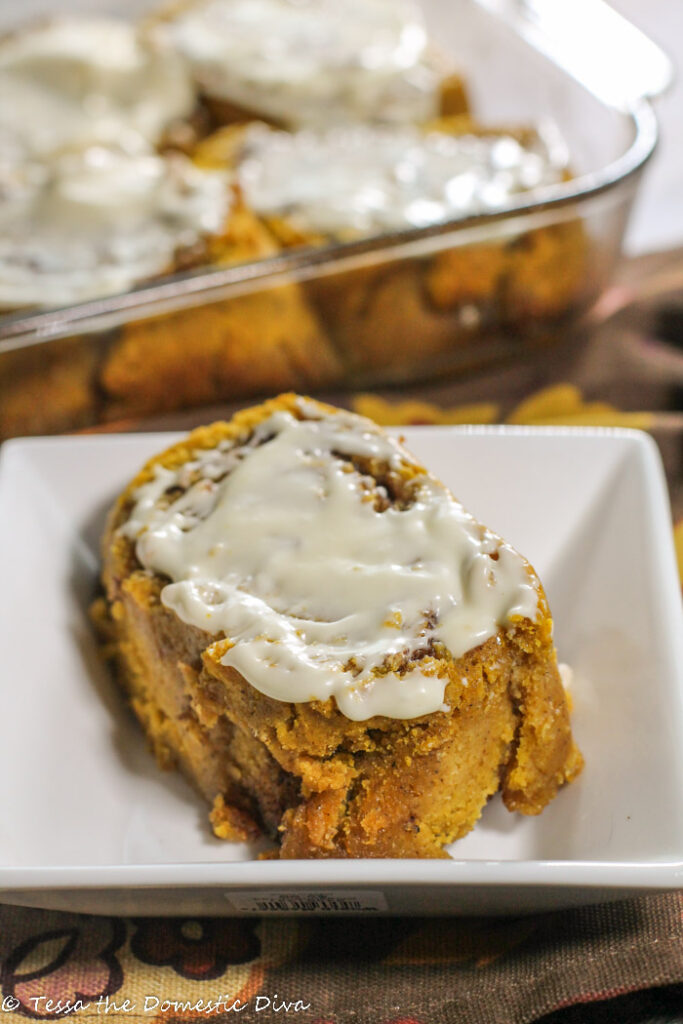 a cream cheese glazed pumpkin cinnamon roll on white plate with the full pan in background