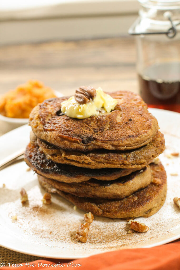 a stack of pumpkin pancakes on a white pate with a dusting of cinnamon and a topped with butter and a whole pecan