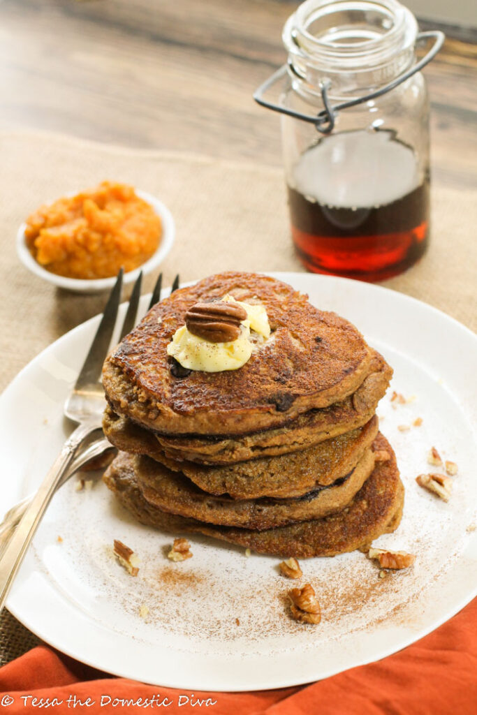 a white plate with a stack of pumpkin pancakes topped with butter and pecans and a real maple syrup