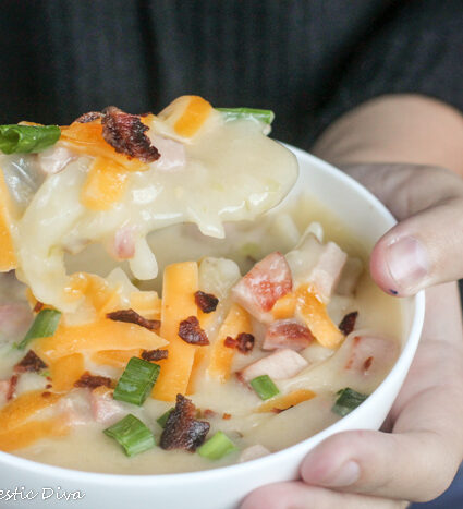 a hand held white bowl with creamy potato soup with cheese, onion, and bacon garnishes