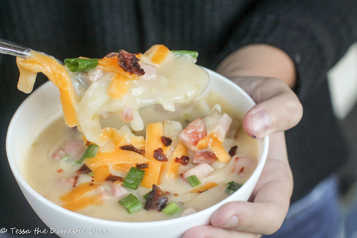 a hand held white bowl with creamy potato soup with cheese, onion, and bacon garnishes