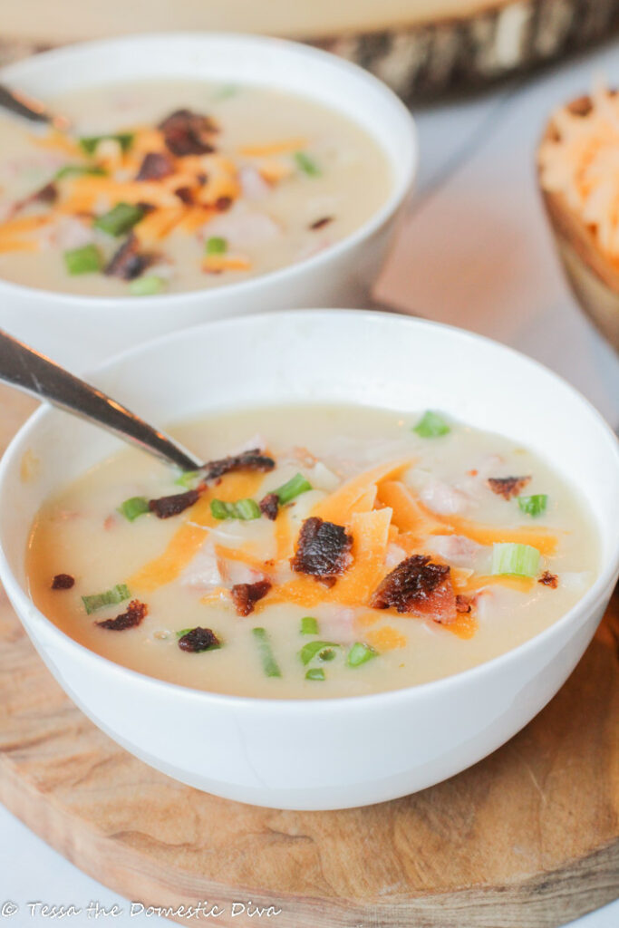 two white bowls atop an olive wood cutting board filled with a a creamy potato soup with cheese, bacon, and green onion garnish