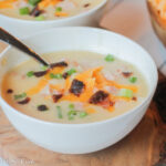 a white bowl on top of a wooden cutting board with a creamy white potato soup with cheddar cheese and chopped bacon topping