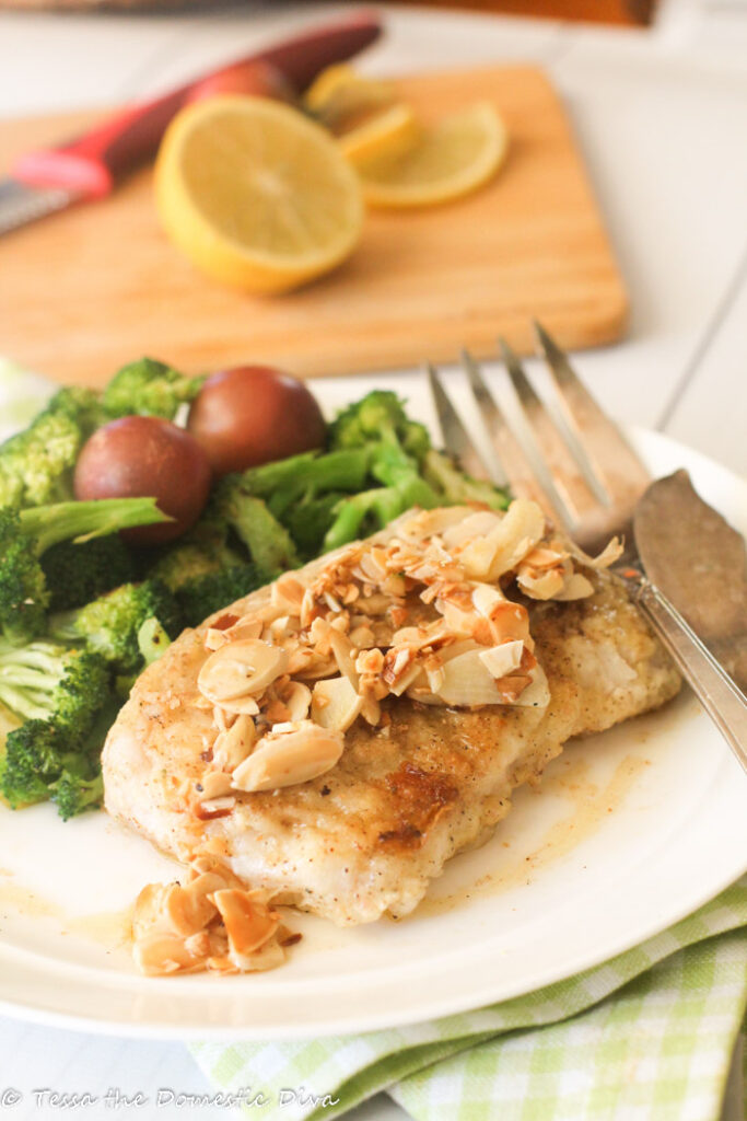 crispy fish on a white plate topped with butter, lemon, and almonds with a side of broccoli