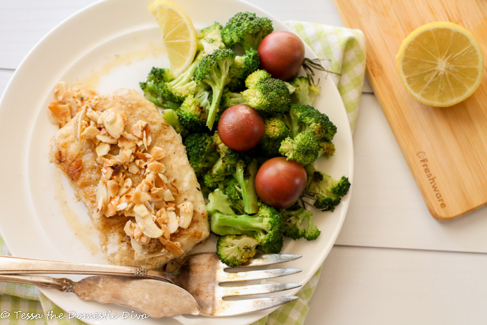 overhead view of a white plate filled with crispy pan friend fish with an almond, garlic, lemon topping and a side of sauteed broccoli and fresh cherry tomatoes