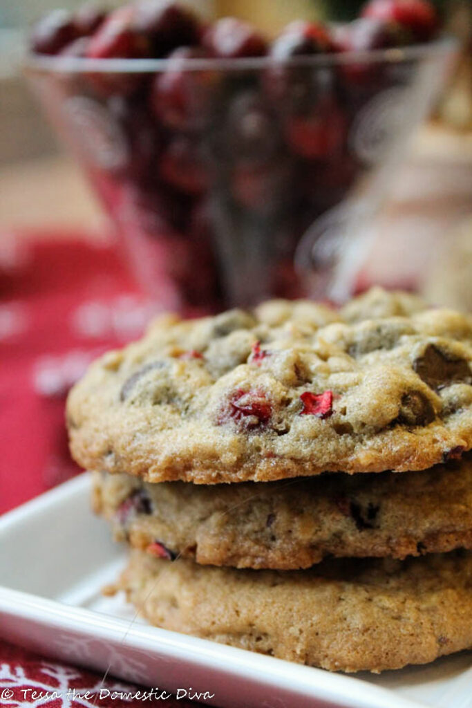 three stacked oatmeal cookies on a square white plate with a cup of fresh cranberries in background