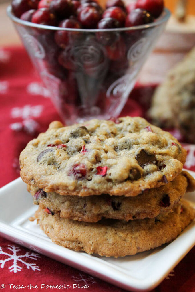 a stack of three oatmeal chocolate chip cookies with bits of chopped fresh cranberries atop a festive red snowflake cloth