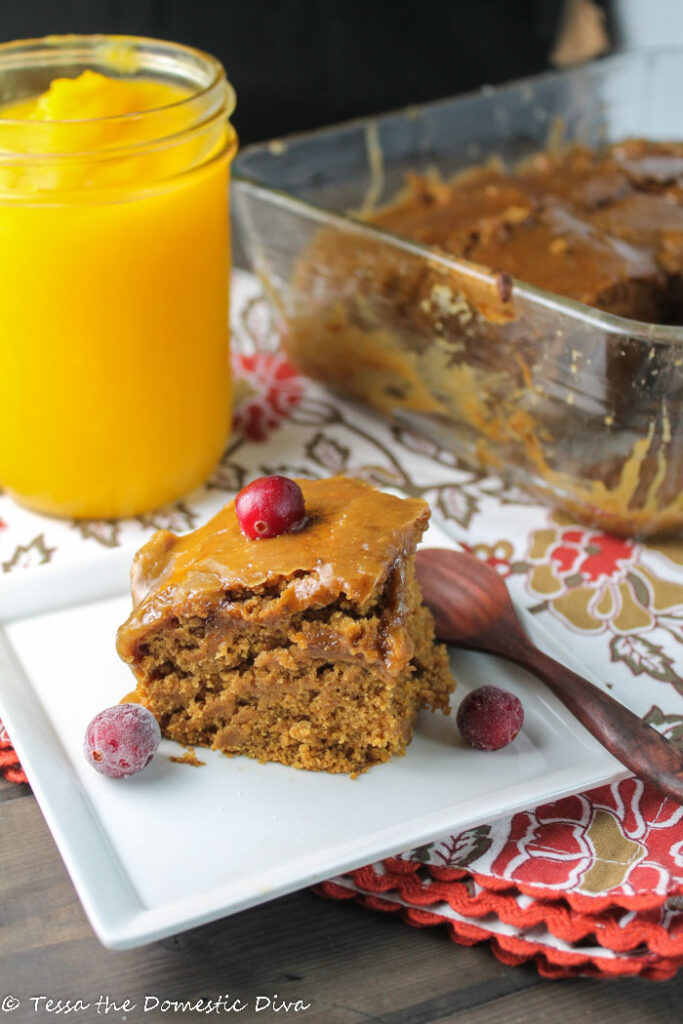 a white square plate with a square of pumpkin cake topped with caramel frosting and a fresh cranberry with a jar of fresh pumpkin puree in the background