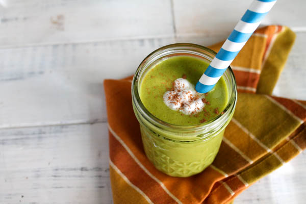 a green pumpkin filled smoothie inside a mason jar with a blue and white striped paper straw atop a autumn colored cloth