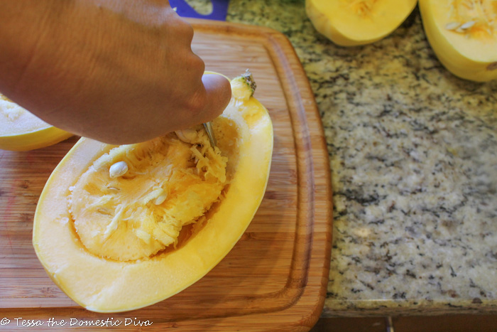 a halved spaghetti squash with the pulp and seeds being scraped out on a wooden cutting board