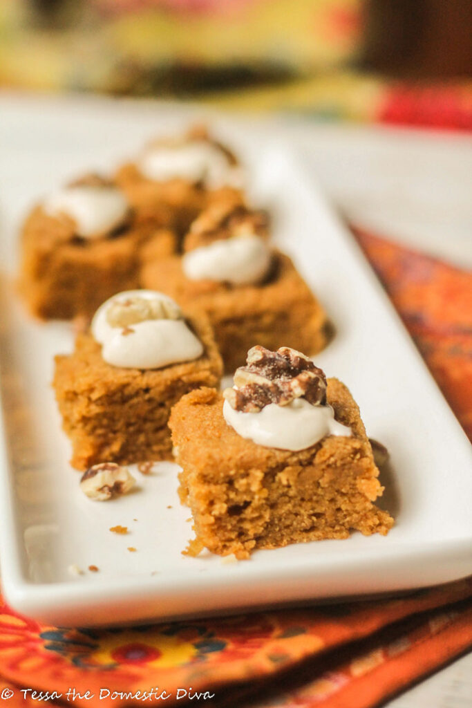 pumpkin cake squares on a white rectangular plate with a dollop of creamy white frosting and a toasted walnut half nestled in the top