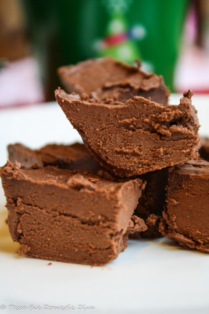 close up of several square of chocolate fudge on a white plate with a holiday green tin in background
