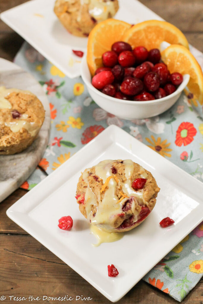 fresh cranberry orange studded muffins topped with an orange glaze and fresh orange zest on a blue floral cloth on white plate