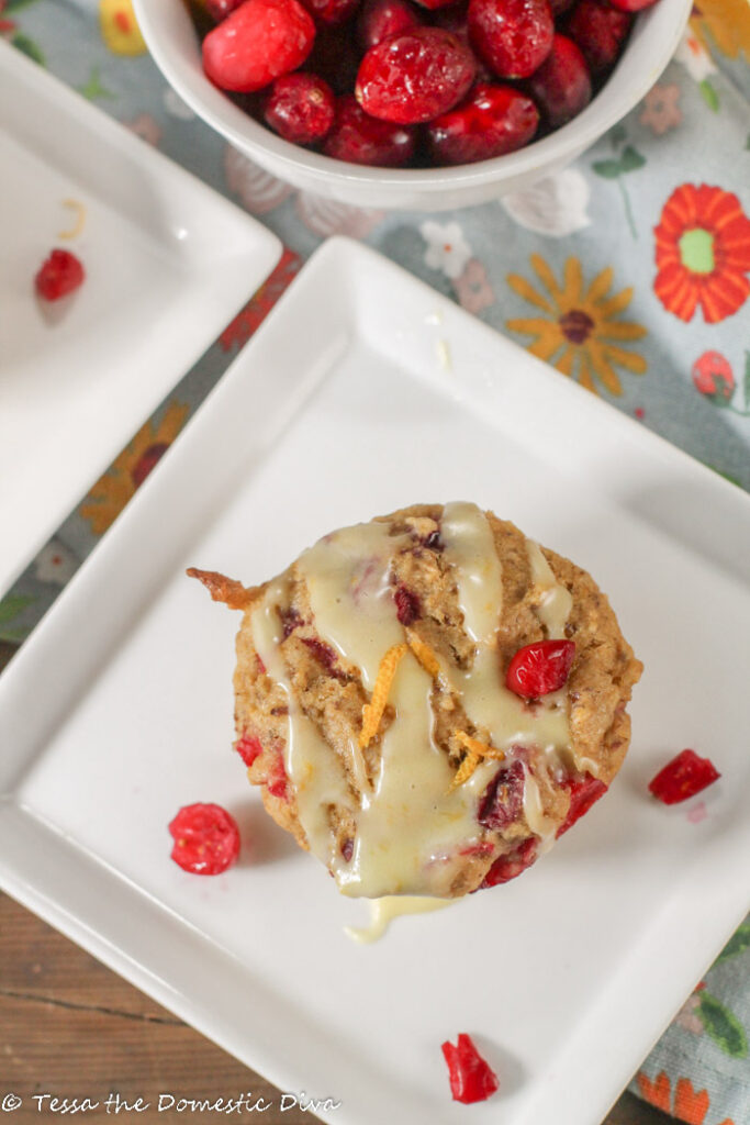 birdseye view of a cranberry orange muffin topped with a drizzle of orange glaze on a white sqaure late with fresh cranberries scattered about