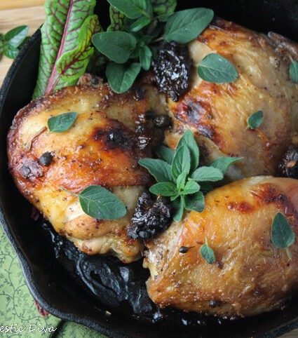 birdseye view of three roasted chicken thighs with crunchy skin in a cast iron pan with chopped prunes and fresh oregano