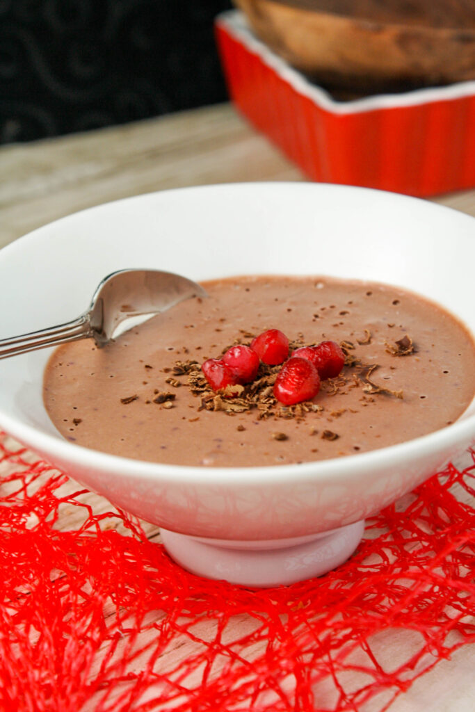 a white bowl filled with a thick chocolate mousse topped with chocolate shavings and pomegranate seeds