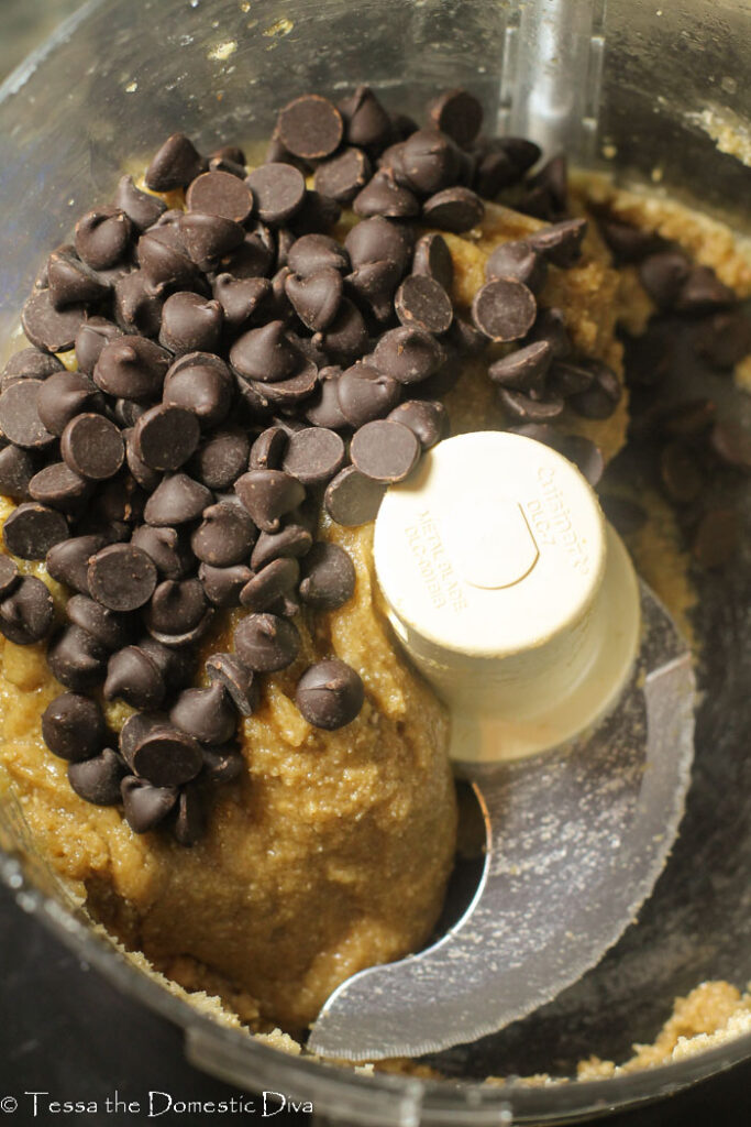 overhead flourless cookie dough batter in a food processor bowl with a pile of chocolate chips on top ready to be mixed in