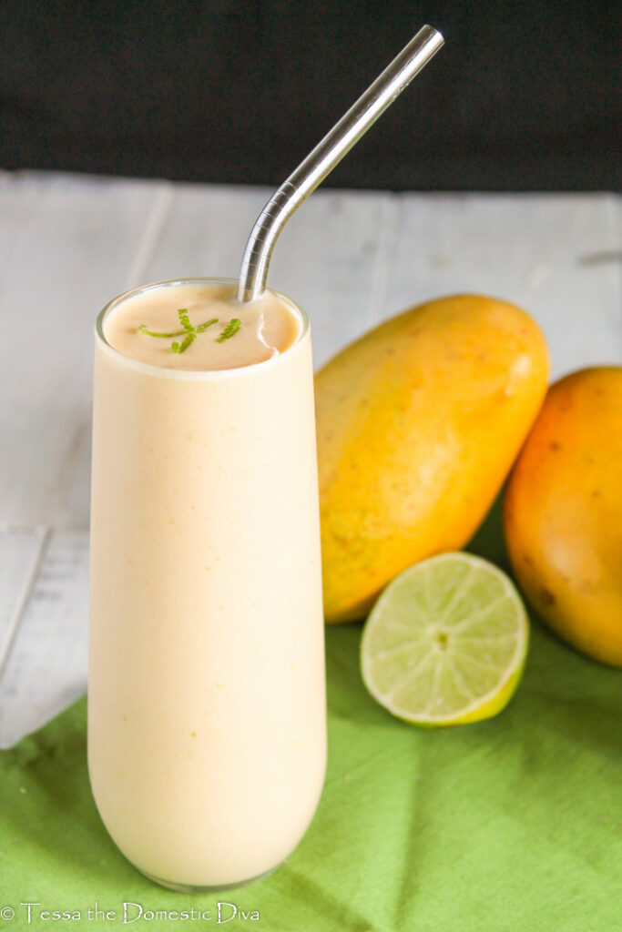 a mango, lime creamy smoothie in a tall glass with a stainless steel straw and fresh lime and Thai mangoes