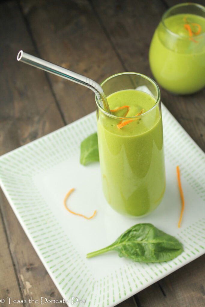 a green smoothie in a clear glass with a stainless steel straw with orange zests and spinach leaves scattered on the white plate