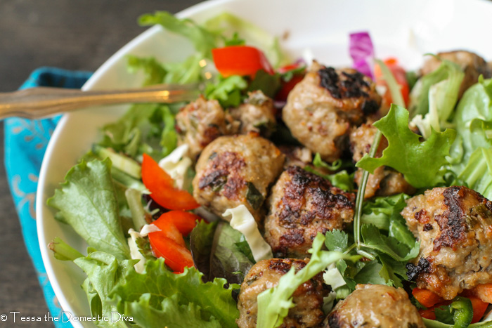 close up a white bowl filled with mixed greens topped with roasted pork meatballs, red peppers, cilantro, and cucumber